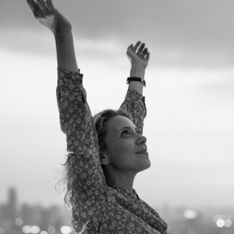 caucasion woman outside raising her arms in the air with a slight smile on your face