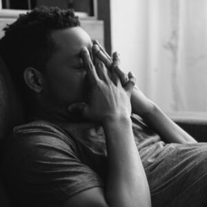 A black man holding his hands to his face in distress with his eyes closed while lying on a bed